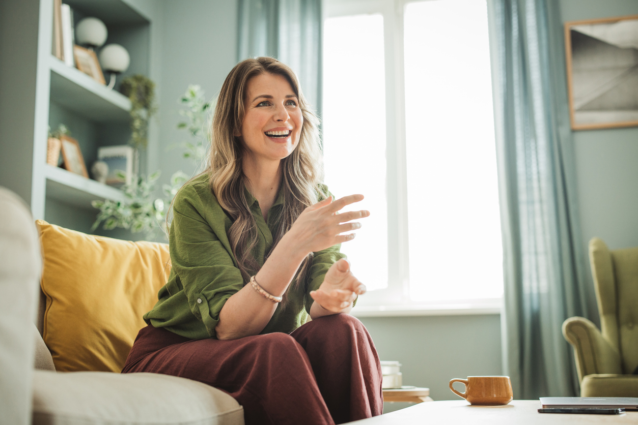 A woman in a green shirt smiles and gestures during a relaxed conversation in a bright, plant-filled living room.