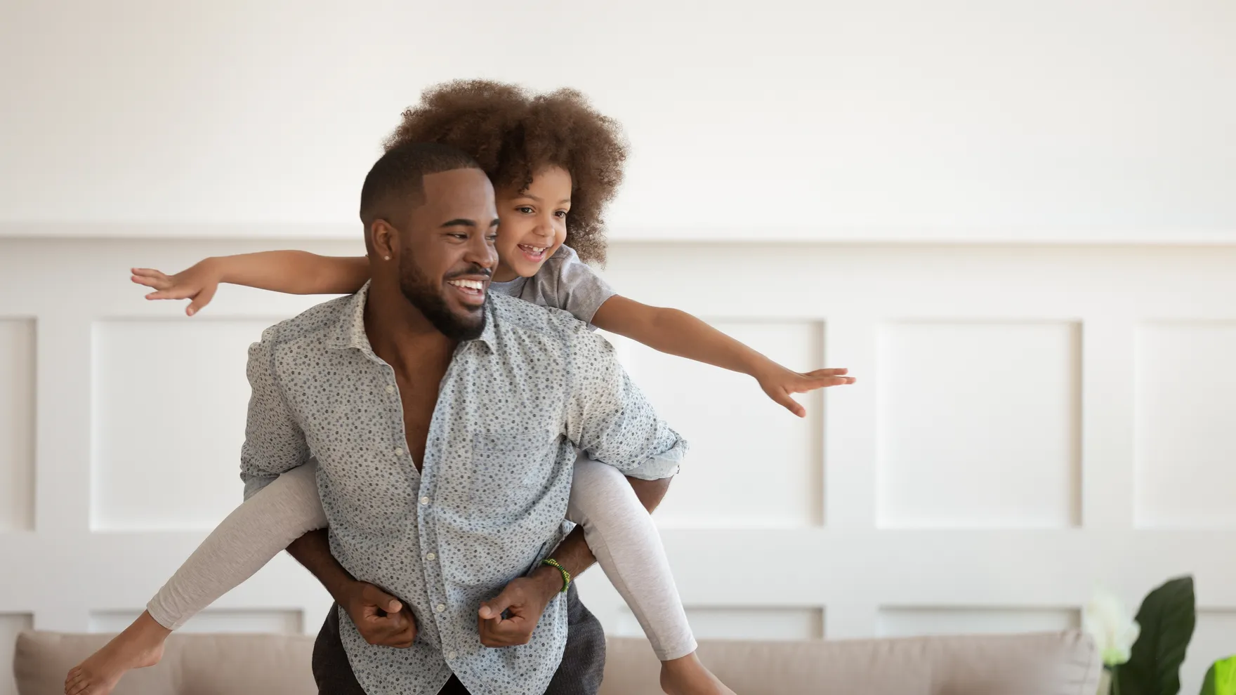 A smiling father giving his laughing young child a piggyback ride at home, arms outstretched like an airplane.